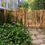 Franse rondhoutpoort van 80 cm bij 80 cm als doorgang naar een tuin met een hondje dat achter de poort zit.