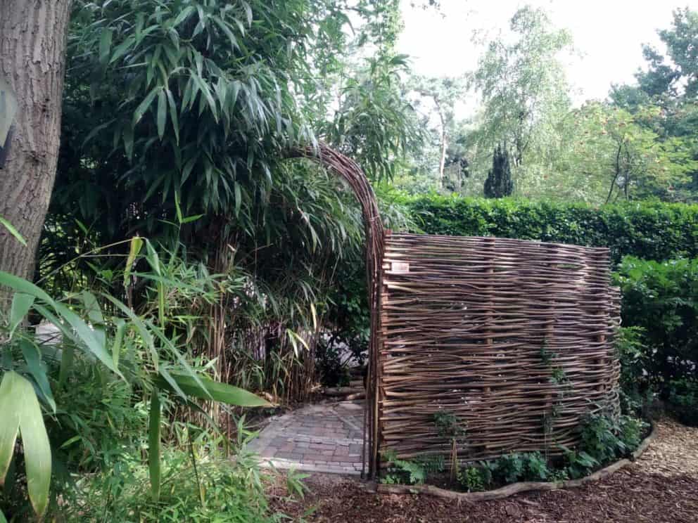 Wilgentenen toegangsboog vastgezet aan een wilgentenen scherm dat in een boog loopt. De wilgentenen staan in de tuin waar veel groen en bomen zijn. Scherm wordt gebruikt als afrastering van een deel in de tuin.