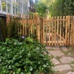 Franse rondhoutpoort van 80 cm bij 80 cm als doorgang naar een tuin met een hondje dat achter de poort zit.