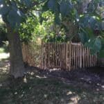 Franse rondhoutpoort van 120 cm hoog en 80 cm breed als doorgang naar een moestuin omheind met schapenhek van 120 cm hoog en een latafstand van 6 cm.