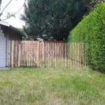 Franse rondhoutpoort van 120 cm hoog bij 100 cm breed als doorgang tussen een heg en een tuinhuisje met schaoenhek van 120 cmhoog en een latafstand van 4 cm ertussen.