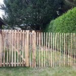 Franse rondhoutpoort van 120 cm hoog bij 100 cm breed als doorgang tussen een heg en een tuinhuisje met schaoenhek van 120 cmhoog en een latafstand van 4 cm ertussen.