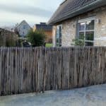 Dubbele Franse op maat gemaakte rondhoutpoort van 114 cm breed bij 120 cm hoog en het andere deel 250 cm breed en 120 cm hoog en afgewerkt met halfronde latten tegen elkaar en bevestigd tussen het huis en de heg.