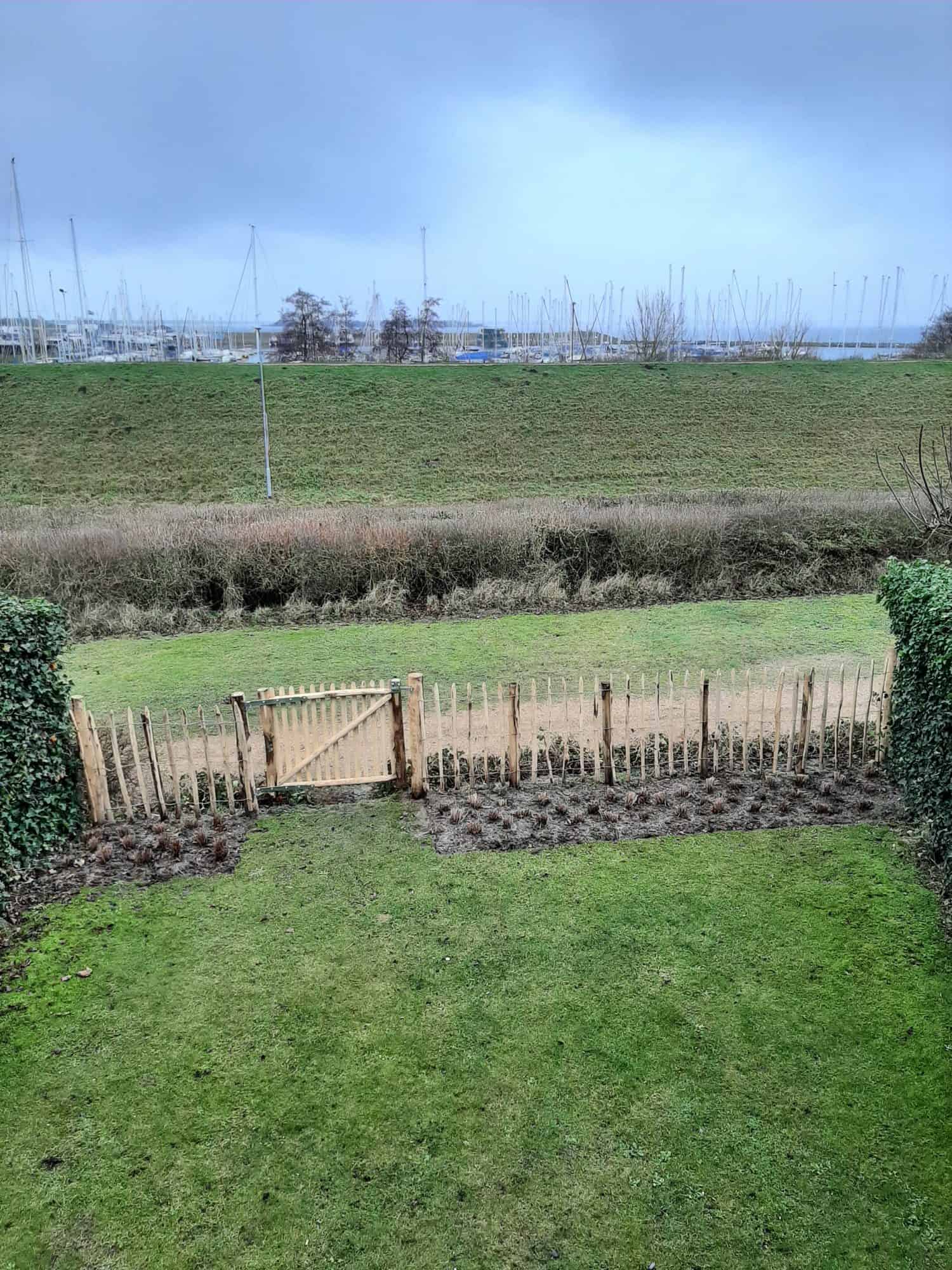 Franse rondhoutpoort van 120 cm breed bij 100 cm hoog tussen schapenhek van 100 cm hoog met een latafstand van 10 cm als afscheiding van een tuin dichtbij de haven.
