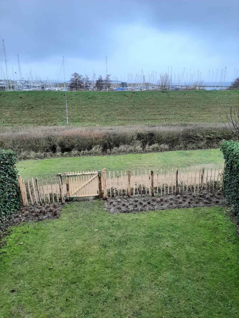 Franse rondhoutpoort van 120 cm breed bij 100 cm hoog tussen schapenhek van 100 cm hoog met een latafstand van 10 cm als afscheiding van een tuin dichtbij de haven.