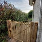 Franse rondhoutpoort van 120 cm breed bij 120 cm hoog geplaatst tussen de zijkant van een woning en een heg.