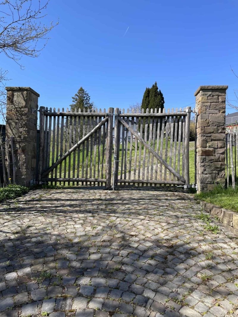 Op de foto is een gesloten rustieke dubbele Franse rondhoutpoort van 150 cm hoog en 138 cm breed van Adequat te zien. De poort is bevestigd aan twee stenen pilaren van ongeveer 200 cm hoog. Naast de pilaren is nog een klein stukje hekwerk zichtbaar. Dit hekwerk is ongeveer 100 cm hoog. Op de foto is de oprijlaan te zien, deze bestaat uit klinkers. Achter de poort kan je vaag wat gras en bomen zien van het woonerf dat zich in Duitsland bevindt.