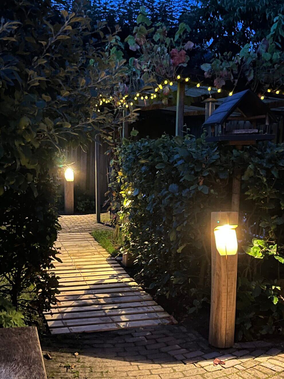 Kastanje looppad met houten op maat gemaakte tuinverlichting Bella in rustiek vierkant formaat, op voeten geplaatst (niet in de grond) met stopcontact outlet aan achterzijde. Kastanje hekwerk horizontaal bevestigd aan schutting.